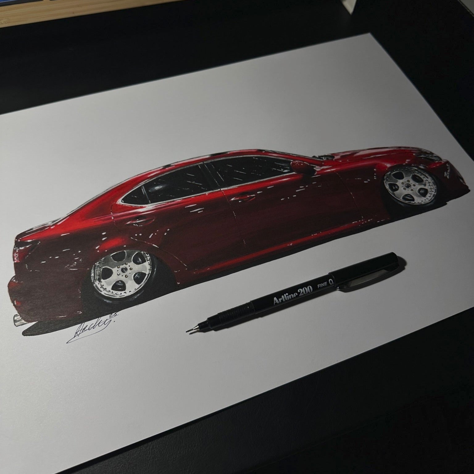 Red car drawing on white paper with a pen next to it, on a desk.