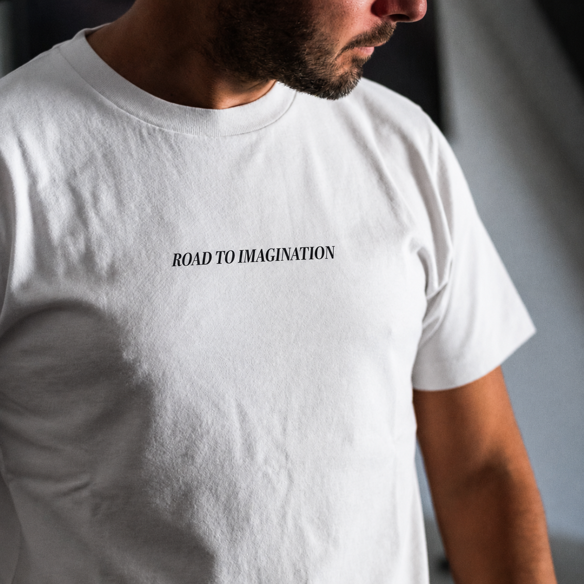 A close-up of a man wearing a white crew-neck t-shirt with the black printed text 'ROAD TO IMAGINATION' on the chest.