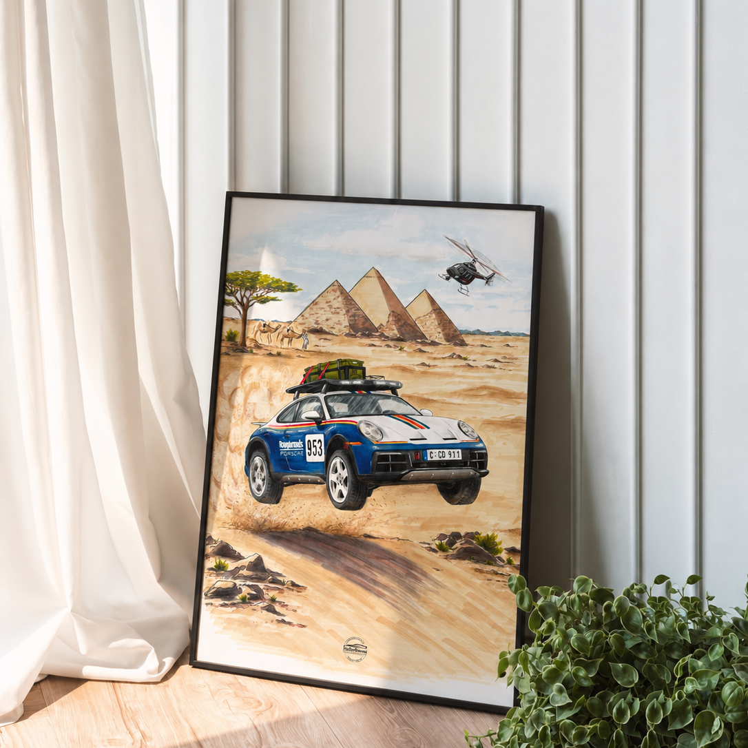 A framed art print leaning against a white paneled wall on a wooden floor, showing a blue Porsche Dakar 911 drawing across a desert toward pyramids with a helicopter overhead. A white curtain and green plant are visible around it.